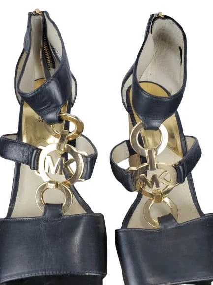 Black high-heeled shoes with gold 'MK' logo on a white background
